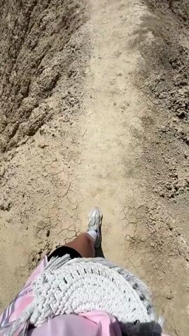 The video shows a person walking along a narrow dirt path atop a ridge in a desert landscape. The path is cracked and dry, and the terrain on either side drops steeply into a canyon.  In the distance, snow-capped mountains are visible under a clear blue sky.  Another person is visible further along the path. The camera perspective is first-person, showing the person's legs and feet as they walk. They are wearing shorts, white socks, and gray sneakers. A white, fringed shawl is visible over their back.