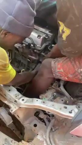 Two people are shown working on a car engine. One person is wearing a yellow shirt and a blue beanie, and the other person is wearing a black shirt and patterned shorts. They are both leaning over the front of the car, reaching into the engine compartment. The car frame is visible, with various parts and wires exposed. The engine is silver and black, and appears to be partially disassembled. The people are working together, their arms and hands intertwined as they manipulate parts of the engine. The environment seems to be an outdoor work area, possibly a repair shop or garage.
