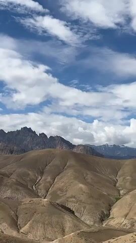 A landscape video shows rolling brown hills with a jagged mountain range in the background under a blue sky with white clouds. A tall, metal power line tower stands on the right side of the frame, and a dirt road winds through the hills. The camera pans from left to right, capturing the expansive scenery.