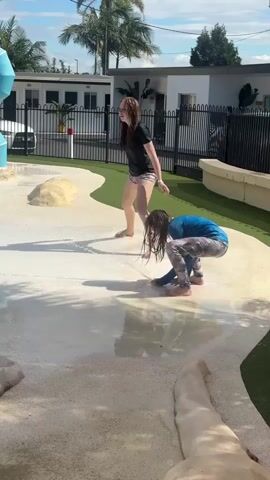 A young person stands on a wet, sandy surface, leaning forward with their arms outstretched. They are wearing a black t-shirt and patterned shorts. Another young person is crouched on the same surface, facing away from the camera, wearing a blue long-sleeved shirt and patterned leggings. Water is spraying from the left side of the frame, creating splashes on the ground and on the individuals. The environment appears to be an outdoor water play area with palm trees and buildings in the background under a partly cloudy sky.