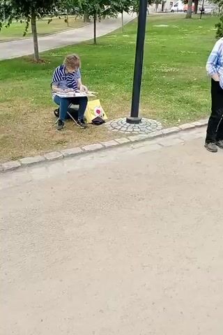 The video shows a park scene with several people, mostly women, sitting on a bench and engaged in artistic activities. In the foreground, a woman is seated on the ground and sketching on a canvas. She is wearing a blue and white striped shirt and dark pants. Beside her is a yellow tote bag. Further down the bench, three women are seated. The woman in the middle is wearing a straw hat, a light-colored jacket, and white pants, and is holding a sketchbook. Next to her, a woman in a colorful scarf and maroon jacket is also looking at a sketchbook. On the far end of the bench, another woman is seated and appears to be working on something on a laptop. A bicycle is parked behind her. The environment is a park with a paved pathway in the foreground, grass, trees, and benches. Another person, a woman, is standing on the left side of the frame, observing. She is wearing a blue and white checkered shirt, black pants, and a white hat. The lighting suggests it is daytime, likely overcast or late afternoon given the soft light.