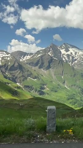 The video shows a scenic view of a mountain range with patches of snow on the peaks. The slopes are covered in lush green grass, and there are some small white flowers scattered throughout. The sky is blue with scattered white clouds. In the foreground, there is a stone marker and a wooden post by the side of a road. The camera appears to be moving slowly, possibly from a vehicle, offering a panning view of the landscape. The lighting suggests it is daytime, with sunlight casting shadows on the mountainside. There are no people or animals visible.