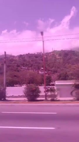 The media file captures a view from a moving vehicle on a road in La Guaira, Venezuela. The scene is characterized by a purple-tinted sky with scattered clouds, suggesting it might be late afternoon or a time with unusual lighting conditions. The landscape features rolling hills covered in vegetation and scattered buildings, indicative of a semi-urban or suburban environment.

Several objects are visible:
- A dark-colored SUV is driving on the road.
- A smaller white vehicle is also seen.
- A lamppost with a circular light fixture is present.
- A building with a white facade and a green roof, featuring a cross on top, is visible on the right side of the road.
- Further down the road, a larger, multi-story building with a light brown facade and numerous windows is prominent.
- Trees line the side of the road in front of these buildings.
- Road markings, including white and yellow lines, are clearly visible.

The activity taking place is vehicular movement along a paved road. The setting appears to be a roadside environment with a backdrop of hills and buildings. There are no clearly discernible people in the footage.

Notable details include the prominent purple hue cast over the entire scene, which could be due to camera settings, filters, or atmospheric conditions. The presence of a building with a cross suggests a potential religious or institutional site. The hilly terrain and the style of architecture hint at a location in a tropical or subtropical region.

There is some visible text on a building to the left of the frame, which appears to be part of a sign with letters like "PER" visible. However, the full text is not legible.

The overall mood is neutral, primarily observational due to the perspective from a moving vehicle. The time of day is not precisely ascertainable but the lighting suggests daylight. The weather appears clear enough for visibility, despite the sky's unusual color.