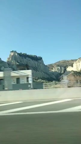 The video shows a view from a moving vehicle on a highway. In the foreground, there is a highway with white markings on the asphalt. A silver barrier separates the highway from the landscape. In the background, there are hills and mountains with sparse vegetation, characterized by rocky slopes and some trees. The sky is clear and blue. A white building is partially visible on the right side of the frame. There is a lamppost on the left side of the frame. The video appears to be filmed during the day.