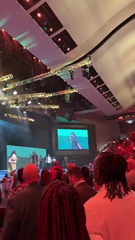 The video captures a live event taking place on a stage in a large indoor venue. The stage is illuminated by numerous spotlights, creating beams of light that cut through the darkness. In the background, a large screen displays a green landscape. On stage, several individuals are present, some appearing to be performers or speakers, and others possibly part of a choir or band. The audience is visible in the foreground, seated or standing, facing the stage. The atmosphere appears to be one of engagement and worship, suggested by the presence of performers and the reverent posture of some audience members. The overall scene is dynamic, with movement and light contributing to the lively ambiance.