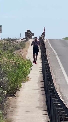 A person is jogging towards the camera on a concrete sidewalk. The person is wearing a dark cap, sunglasses, a patterned shirt with dark sleeves, dark shorts, and bright pink running shoes. The sidewalk is adjacent to a road, separated by a metal barrier. Beyond the sidewalk, there is green shrubbery. In the distance, the road slopes upwards, ending at a barrier with a sign. The sky is clear and bright. The overall environment appears to be sunny and outdoors.