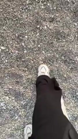 The video opens with a low-angle shot of a person's feet walking on a gravelly surface, before panning up to reveal a beach scene. The person is wearing black pants with a white stripe and white sneakers. The beach consists of small, round pebbles. The camera then shifts to show the ocean, with small waves lapping against the shore. In the background, there is a distant coastline and a partly cloudy sky with the sun shining through. The overall lighting suggests either sunrise or sunset.