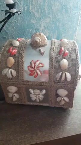 The video shows a close-up of a decorative chest or box. The chest is covered in burlap and adorned with various seashells, including some red-colored ones and larger, more prominent shells at the top and front. There are also some decorative painted elements on the chest, resembling flowers or starbursts. The chest sits on a wooden surface, possibly a table. The background appears to be a textured wall, possibly with a blue or green hue. The lighting is somewhat dim, and the camera angle provides a stationary view of the object, with slight panning and zooming movements. There are no people visible in the video.
