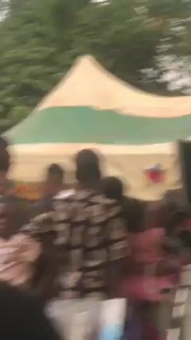 The video shows a gathering of people under a tent. The tent has green and white stripes. A man in a striped tank top walks past a sound system. Other people are dressed in various outfits. Some people are holding children. Trees are visible in the background. The scene seems to be outdoors.