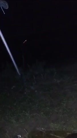The video shows a dark outdoor scene at night. A bright light, possibly from a flashlight or camera, illuminates a patch of grass and some vegetation. In the distance, there is a faint light source, possibly a sparkler or a small fire. There are no people or animals visible in the frame. The ground is covered with grass and dirt. There is a white pole-like object on the left side of the frame.