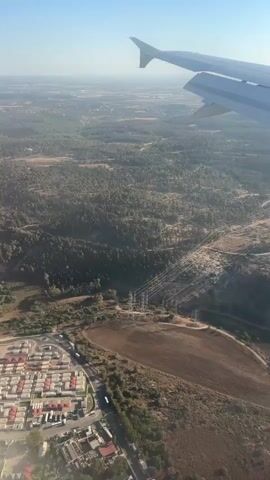 This aerial view captures a landscape from an airplane window, showcasing a combination of natural and man-made features. The primary subjects are the vast expanse of green trees, a winding path through the terrain, a residential area with red-tiled roofs, and a prominent electrical transmission line with tall towers and wires. The scene is set in a rural or semi-rural area, likely Beit Nehemia, Israel, given the location context.

The activity is implied to be flight, with the airplane's wing visible in the upper right corner, suggesting movement through the sky. The time of day appears to be daytime, with clear skies and bright sunlight casting shadows, indicating fair weather. The landscape is characterized by rolling hills covered in dense forestation, interspersed with cleared areas that are likely agricultural fields. The residential area shows a grid-like structure of houses and streets, suggesting a small town or suburban development. The electrical transmission line is a significant feature, cutting through the landscape and indicating the presence of infrastructure.

Notable details include the contrast between the natural forest and the built environment. The shadows suggest the sun is at an angle, possibly late morning or early afternoon. The texture of the agricultural fields and the dense canopy of the trees are visible. The presence of the airplane wing signifies the perspective and the mode of observation. No visible text is present in the media file. The overall impression is one of a journey over a varied and developed landscape.