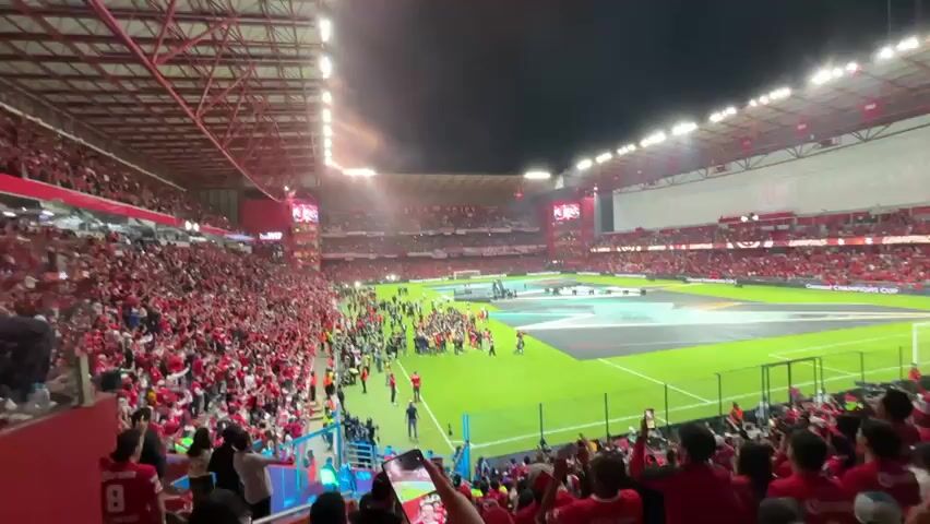 The video shows a soccer stadium filled with a large crowd of fans mostly dressed in red. They are cheering and celebrating. The field is green and has several people on it, likely players and staff. The stadium is covered and well-lit. There are large screens visible in the stadium. Some people in the crowd are holding up their phones, presumably to record the event. The atmosphere is lively and celebratory.