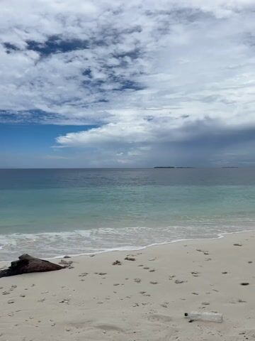 The video showcases a tranquil beach scene in Ukulhas, Maldives, transitioning from sunny to overcast conditions. Initially, a wide shot captures a pristine white sandy beach with gentle waves lapping at the shore. The clear turquoise ocean stretches to the horizon under a bright blue sky dotted with fluffy white clouds. Pieces of dark driftwood and coral fragments are scattered on the sand, with a plastic bottle briefly visible on the bottom right.

As the video progresses, the camera pans across the coastline. A boat is seen in the distance on the ocean. The sky gradually becomes more overcast, with grey clouds beginning to dominate. The scene then focuses on a person standing on the sandy beach, actively engaged in fishing. They are holding a long fishing rod and casting their line into the ocean, facing away from the camera. The sound of gentle waves breaking on the shore is audible throughout the clip.