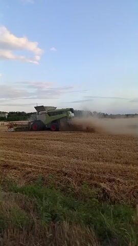 A combine harvester is working in a field of cut crops, kicking up dust as it operates. The sky above is blue with scattered clouds. The field appears to be recently harvested, with stubble visible. In the background, trees and possibly buildings can be seen. The combine is green and white with red wheels.