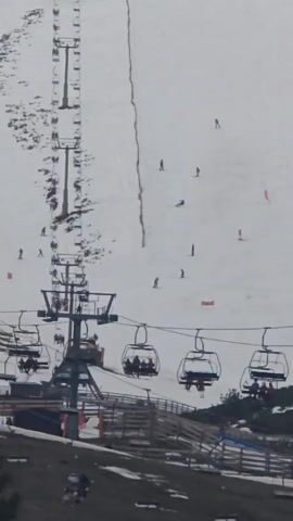 The video shows a ski slope with a chairlift. Several people are skiing down the slope. The sky is overcast. The chairs on the lift are moving, some with people seated in them. A wooden fence is visible below the lift, and patches of bare earth are visible outside the snow-covered area.
