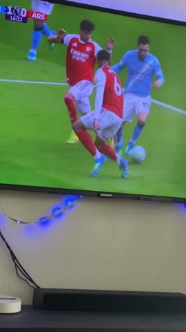 Here is a detailed description of the video:

The video shows a person filming a television screen. The television is displaying a soccer match between Manchester City and Arsenal. The score is 1-0 in favor of Manchester City. A Manchester City player with the number 10 on his back dribbles the ball through several Arsenal defenders. The video is taken from a position in front of and below the television. A soundbar is visible below the TV, and a small white device and some cables are visible on the shelf to the left of the soundbar. A string of blue LED lights is also visible behind the television.