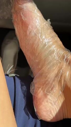 A close-up shot shows a person's lower leg and foot, wrapped in clear plastic wrap. The shot zooms out to show the person's hand holding a small black tube of "NO TATTOO PAIN NUMBING CREAM" with green text, and a small black bottle of "NO TATTOO PAIN OFFICIAL 50% EXTRA NUMB PROCEDURE NUMBING SPRAY-QUICK!" with yellow text. The person is wearing blue bottoms. The lighting appears to be natural daylight.