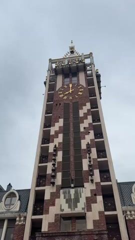 The video shows a tall clock tower building with a large golden clock face. The building is made of brown brick and has a pattern of lighter-colored bricks on its facade. There are small balconies on each level of the tower. The video pans down to show a mosaic on the ground in front of the building. The mosaic is colorful and appears to depict people and other figures. The ground is wet, suggesting it has recently rained. Other buildings are visible in the background. The sky is overcast. The bells on the tower are sounding.