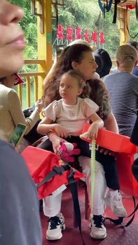 The video shows a woman holding a young girl on her lap on what appears to be a boat. The girl is holding a pink stuffed unicorn and a green toy. They are surrounded by other people, and there are red life vests nearby. The boat has a wooden structure with decorations. The background shows greenery, suggesting they are on a river or lake surrounded by trees. The camera perspective is from someone standing or sitting nearby.