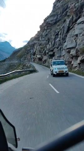 The video shows a winding road carved into the side of a steep, rocky mountain. The camera appears to be mounted on a vehicle, possibly a motorcycle, as indicated by the handlebars visible in the foreground. The road has a white dashed line marking the center. To the left of the road, there is a metal guardrail and a steep drop-off. To the right, the imposing rock face of the mountain rises sharply. In the distance, on the right side of the road, a white vehicle, possibly a van or camper, is partially visible, moving in the same direction as the camera. The sky is overcast, suggesting a cloudy or hazy day. The overall environment is mountainous and rugged, with evidence of human infrastructure in the form of the road and guardrail.