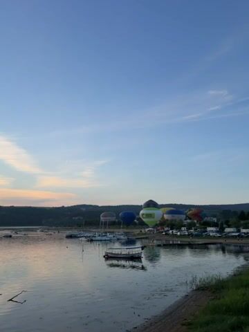A wide shot shows a calm body of water with several boats docked along the shore. In the background, multiple hot air balloons of various colors are inflated and on display, some appearing to be prepared for flight. The sky is a clear blue with wispy clouds, and the sun is setting, casting a warm glow over the horizon. The overall atmosphere is serene and festive.