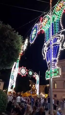 The video captures an outdoor nighttime scene illuminated by elaborate, colorful light displays suspended across a street. The lights are arranged in large arched structures and spherical ornaments, featuring a vibrant palette of red, green, blue, and white LEDs. These light installations are strung between tall poles and possibly buildings, creating a festive atmosphere. Below the lights, a crowd of people is visible, gathered on what appears to be a street or plaza. Some individuals are walking, while others are standing, suggesting a public event or festival. In the background, buildings with illuminated windows can be seen, hinting at an urban or town setting. The overall environment is dark, with the bright artificial lights dominating the visual field.