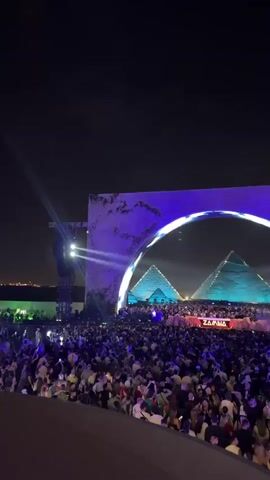 At night, a large crowd is gathered in front of a stage with a large architectural set piece, perhaps a DJ or concert stage. The scene is heavily lit with bright blue lights. The set piece appears to be made of rough blocks, possibly stone. The stage has what appear to be pyramid-like structures on it. The sky is dark, and other lights are seen in the distance. The people in the crowd are moving and appear to be facing the stage, possibly dancing. The name “Zazara” is visible on the stage.