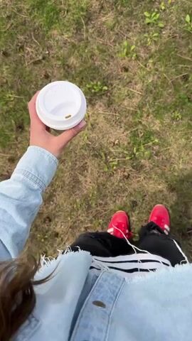 A person walks across grass holding a closed white paper cup. The person is wearing a light blue denim jacket, black ripped jeans, and red sneakers with white stripes. The ground is covered in grass, with some bare patches and small pine cones. The camera's point of view is from above, looking down at the person walking.