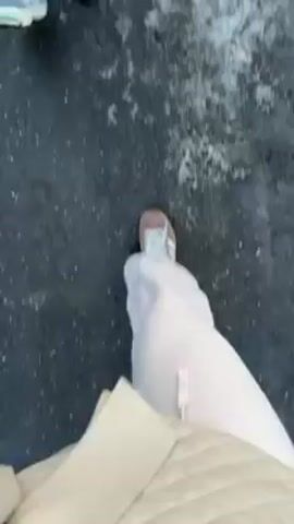 The video shows a person's-eye-view of two pairs of feet walking on a street. The person recording is wearing off-white pants and beige shoes with what appear to be clear plastic shoe covers. Another person, only their feet visible, is wearing white and yellow sneakers. The street they are walking on is asphalt, which is wet with puddles of water and some patches of ice. Traffic noise is audible in the background.