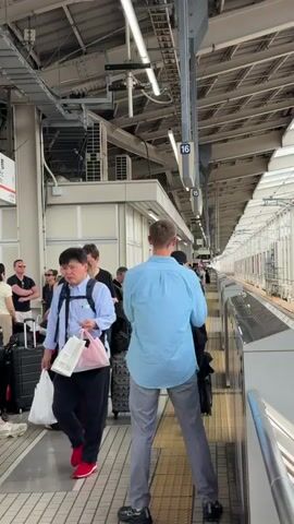 The video shows a busy train platform. Multiple people are waiting, some with luggage, suggesting travel. The people are diverse in age and ethnicity, dressed in casual clothing. One man in a blue shirt and grey pants stands facing away from the camera, holding a phone up as if taking a picture or video. Another man in a black suit and hat is closer to the camera, also with luggage. The platform is covered by a roof with industrial lighting. Tracks are visible to the right, with a train likely approaching or departing. The overall environment suggests a public transportation hub in an urban setting, possibly in Japan given the signage.