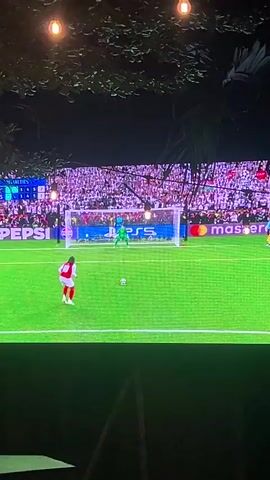 A person watches a soccer penalty kick on television. The player in white and red kicks the ball to the left side of the goalie, scoring. The room is dark, illuminated only by the television light, with a person in the foreground and plantlife and some small hanging lights visible at the top of the frame.