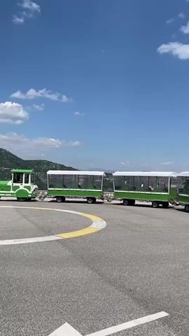 The media file depicts a bright, sunny day in Lipa Dobrska, Montenegro. The scene features a tourist train with several green carriages with white canopies, parked on an asphalt surface marked with white lines. In the background, there's a building with a green roof and a railing, suggesting a public area or a tourist attraction. Several people are visible in the background, some standing near cars and others walking. Two white cars are parked near the building. The sky is a clear blue with scattered white clouds, indicating pleasant weather. The overall impression is that of a tourist destination on a beautiful summer day, with visitors arriving and departing. There is a license plate visible on one of the white cars with the text "PG LT ".