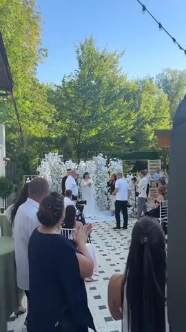 A wedding ceremony is taking place outdoors in daylight. A bride in a white dress and groom in a black suit are standing at an altar adorned with white flowers. A man in a white t-shirt is standing near them, appearing to be a photographer or officiant, holding a camera. Several guests are seated in the foreground, facing the altar and clapping. Others are standing around the periphery. Trees and foliage are visible in the background.