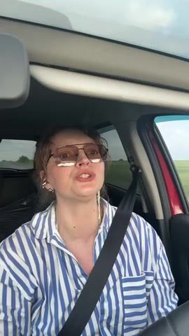 A medium shot shows a white woman with short brown hair, wearing sunglasses and a blue and white striped shirt, sitting in the driver's seat of a car. She is looking to the right of the frame and appears to be singing or talking. A black seatbelt is visible across her chest. The background shows a blurry green landscape and a cloudy sky.