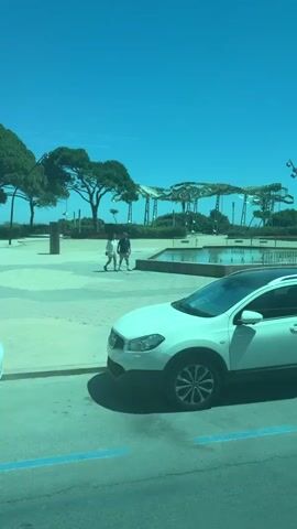 The video captures a view from a moving vehicle, likely a bus or car, looking out at an urban waterfront area on a bright, sunny day with a clear blue sky. In the foreground, a white vehicle is parked in a designated spot marked with blue lines, partially obscuring a pedestrian crossing. Adjacent to it, another car is visible, followed by a silver coupe parked on the street. Further ahead, a man walks on a paved area. The middle ground features a rectangular water fountain with several jets spraying water. Beyond the fountain, a row of distinctive, umbrella-shaped structures rise above a lush green landscape dotted with trees, including several prominent palm trees. Several lamp posts and a bus stop sign are also present. Two motorcycles are parked near a crosswalk, and a white van is parked on the street. The overall impression is a well-maintained public space with architectural features, greenery, and evidence of vehicular and pedestrian activity.