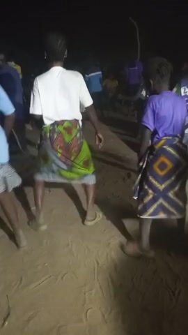 Two people are dancing in an open, sandy area at night. They are both facing away from the camera. The person on the left is wearing a white t-shirt and a colorful printed cloth wrapped around their waist like a skirt. They have dark hair styled in braids and are wearing flat sandals. The person on the right is wearing a purple t-shirt and a different colorful printed cloth wrapped around their waist in a similar fashion. They also have dark hair in braids and are wearing sandals. They are moving their feet and bodies to the music, which has a clear beat. Other people can be seen in the background, but they are out of focus. The lighting is dim.
