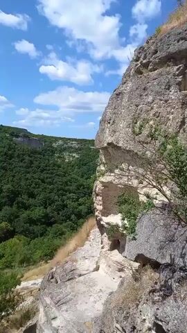The video captures a panoramic view of a lush green forested valley on a sunny day. The sky is bright blue with scattered white clouds. In the foreground, a rugged, rocky cliff face dominates the right side of the frame, with patches of green vegetation growing from its crevices. The cliff appears to be made of light-colored stone, showing layers and erosion. A narrow path or dirt road winds through the grassy slopes below the cliff. The dense forest stretches across the valley, forming a thick canopy of green trees. The overall scene depicts a natural, wild landscape.