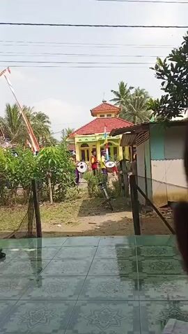 A group of children in yellow and purple uniforms with hats are marching and playing drums in front of a building with a red roof and a dome. There are trees and flags around the building, and the ground is dirt. The video is shot from a distance, through a window.