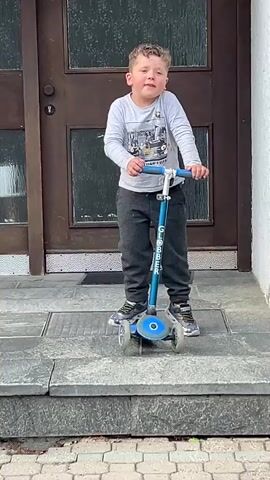 A young boy with curly brown hair, wearing a light gray long-sleeved shirt, dark gray sweatpants, and sneakers, stands on a blue three-wheeled scooter on a light gray tiled step. He holds the scooter handlebars and speaks, gesturing briefly with his right hand. Behind him is a dark brown double door with glass panels, and a light-colored building wall is to the right. A brick pathway is visible at the bottom in front of the step.