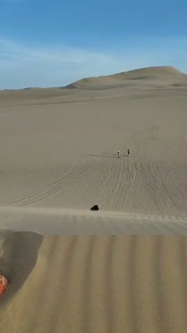 The video shows a wide shot of a desert landscape with a clear blue sky and light clouds above. In the foreground, there are several sand dunes, and in the distance, larger dunes rise higher. The sandy terrain is marked with vehicle tracks and footprints. Three people are visible amidst the dunes. One is lying on their stomach, sliding down a sandy slope, while the other two are standing nearby, watching.