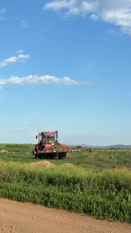 A red tractor with a large attachment is shown in a green field under a blue sky with white clouds. The tractor is moving from left to right across the frame, and the attachment behind it appears to be spreading something on the ground, possibly fertilizer or manure. The field is covered in green grass and some dry, straw-like material. In the background, there are rolling hills. The scene is well-lit by sunlight.