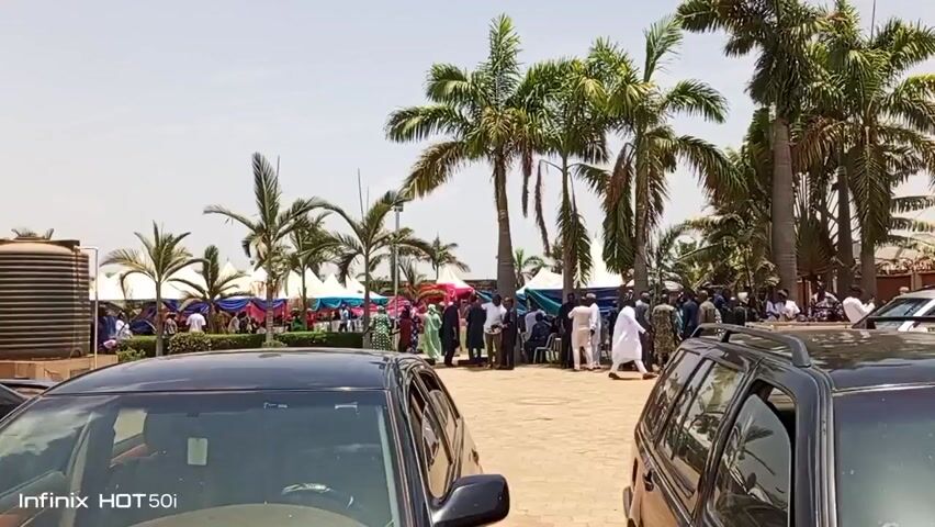 The video depicts a gathering of people outdoors on a paved area. In the foreground, two dark-colored cars are parked, partially obscuring the view. Behind the cars, palm trees are scattered around the area. In the middle distance, a group of people is gathered, some standing and talking, others seated. There are tents or canopies set up, adorned with colorful decorations. In the background, there is a tall, dark water tank. The scene appears to be a daytime event, possibly a social gathering or celebration. The weather seems sunny and clear.