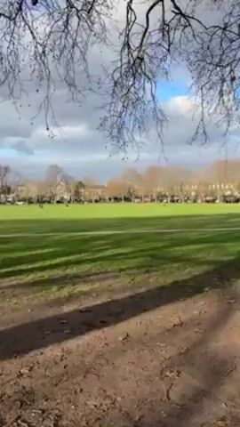 The video shows a wide shot of a large, green grassy field bordered by trees and buildings in the distance. The foreground features a dirt path covered with leaves, partially obscured by the branches of a bare tree. People are scattered across the field, appearing small in the distance. The sky is partly cloudy, with patches of blue visible. The overall scene suggests a public park or common area on a day with mixed sunlight and cloud cover.