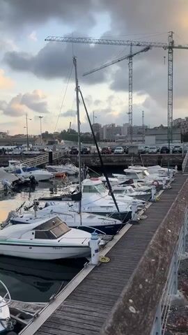 The video shows a marina with several boats docked. The sky is cloudy with some patches of sunlight. There are cranes in the background, suggesting construction or port activity. The boats are of various sizes and types, including small motorboats and a sailboat. The water in the marina appears calm. The video pans across the scene, offering a view of the docked boats and the surrounding environment. No people are clearly visible in the video.