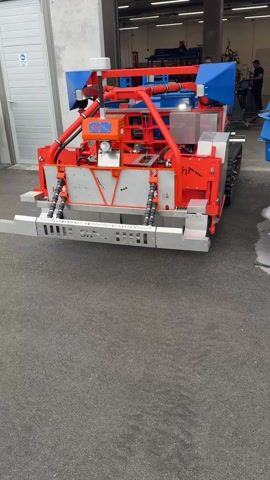 A machine is moving backwards on an asphalt surface towards a building with a garage door. The machine is red, white, and blue, with tank treads visible on its sides. A man is working inside the building in the background. The text "autonomous agricultural system" is written on the front of the machine. There is a satellite receiver on top of the machine.