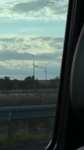 The video captures a view from inside a moving vehicle, looking out at a rural landscape. In the foreground, a hand holding a dark object is partially visible on the left side of the frame. A metal guardrail runs horizontally across the lower portion of the image. Beyond the guardrail, dry grass and sparse vegetation are visible. Two tall lampposts stand in the middle ground, each with several birds perched on the arms and wires. The lampposts have black and white striped bases. In the background, a vast, open field stretches to the horizon under a cloudy sky. The clouds are dramatic, with sunlight breaking through in some areas, casting rays across the sky. The overall scene suggests a drive through a somewhat arid, open countryside during the daytime.