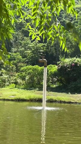 The video shows a large faucet sculpture positioned in the middle of a body of water, with water continuously flowing from it into the lake. The faucet is gold-colored and appears to be made of metal. The surrounding environment is lush and green, with trees and foliage visible in the background. Sunlight filters through the leaves of a tree in the foreground, casting dappled light on the scene. The water from the faucet creates ripples on the surface of the lake. There are no people visible in the video. The overall impression is one of nature and art combined.