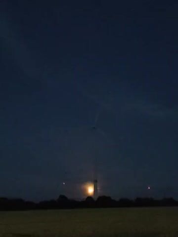 A time-lapse video captures a wind turbine at night. The turbine is tall and white, with three blades visible against a dark blue sky. A bright, yellowish-orange moon is partially obscured by the turbine's base. The foreground features a silhouette of trees and a field of dark, textured grass or crops. The scene is dimly lit, with a few red lights visible on the turbine and on what appear to be power lines in the distance.