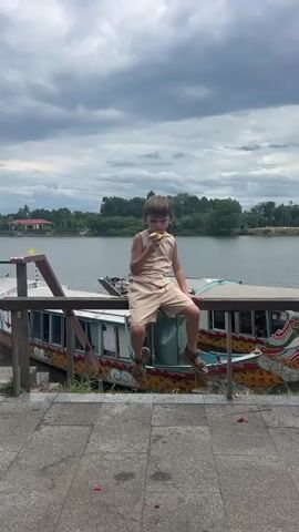A young boy in a tan outfit sits on a wooden railing, eating something yellow. He is facing away from the camera, looking to his left. Behind him is a body of water, with two boats docked on the shore. The sky is cloudy and overcast.