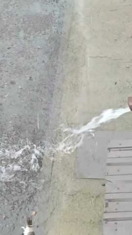 A static overhead shot shows rain falling on a paved surface. The rain is visible as streaks and small splashes on the ground, indicating it is a light to moderate rain. The surface appears to be a mix of asphalt and concrete with some small stones and debris. In the background, a concrete wall with barbed wire on top runs along the edge of the paved area. Beyond the wall, industrial buildings with blue and beige exteriors are visible, along with a large blue crane or gantry. A downspout on the right side of the frame is seen with water spraying out of it, suggesting the rain is causing runoff. There are no people visible in the video.